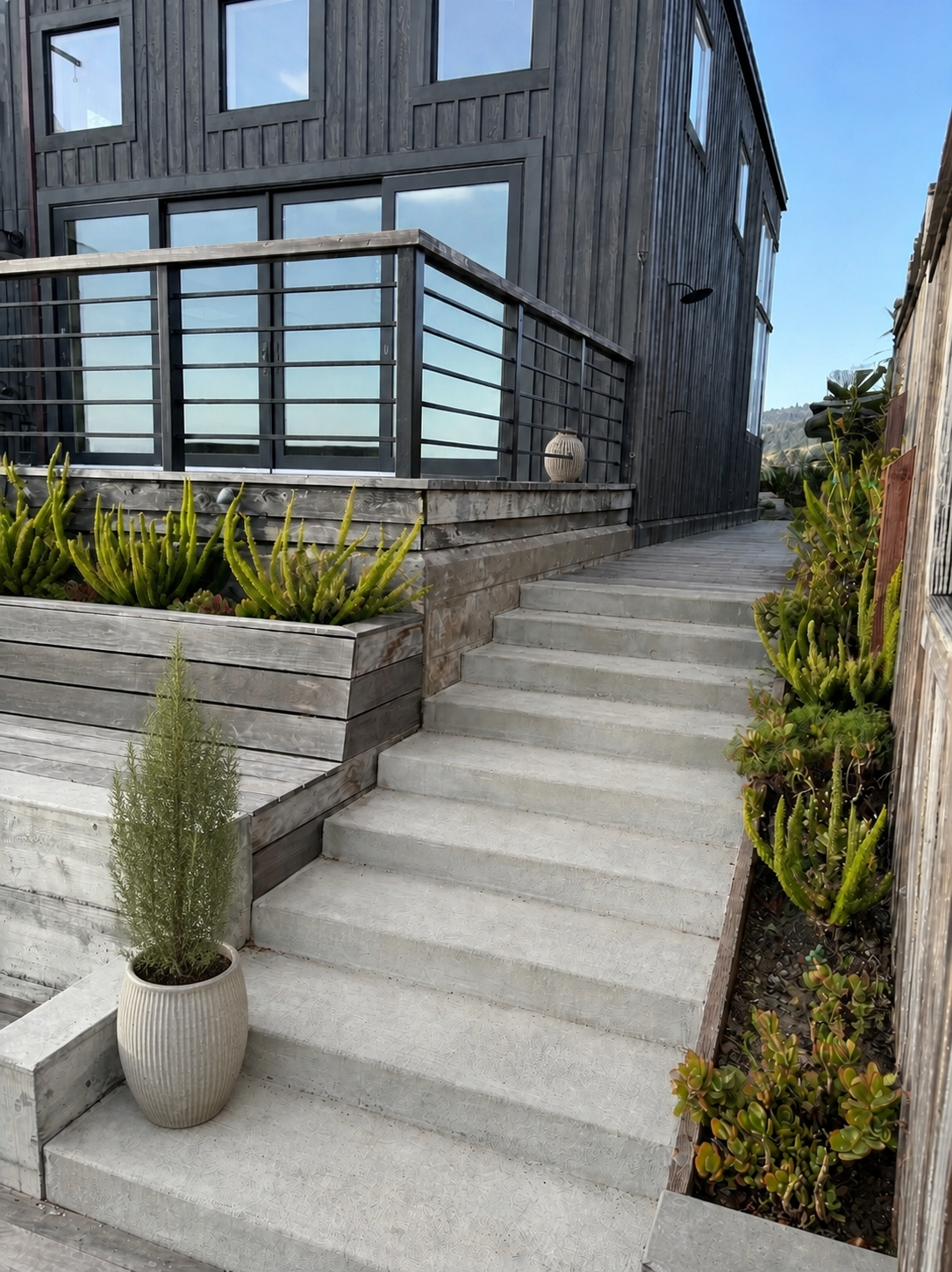 Concrete entry stairs beside a dark wood-clad home with raised planting beds and cable railing